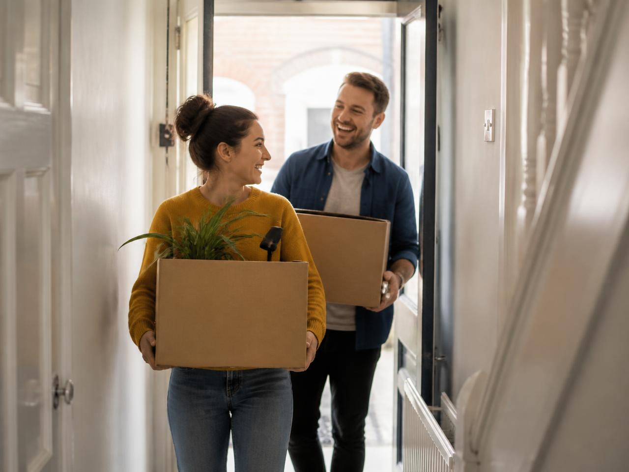 A couple carrying cardboard moving boxes through the hallway of their new home, smiling at each other